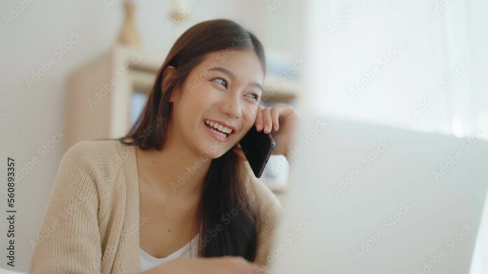 Young asian woman talking mobile smartphone while working on computer laptop on desk in living room at home Young asian woman talking mobile smartphone while working on computer laptop on desk in living room at home