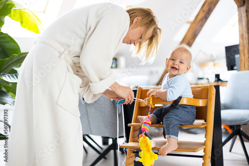 Happy infant sitting and playing with his toy in traditional scandinavian designer wooden high chair in modern bright atic home suppervised by his mother photo