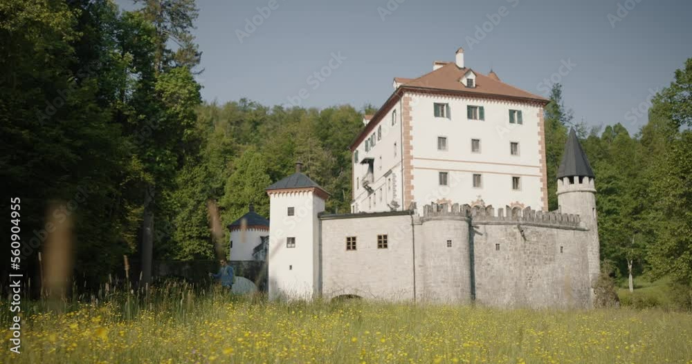 Hiker walking infront of beautiful castle Snežnik in the middle of a spruce forest. Yellow flowers blooming infront on a grassy meadow. Clear sky. Hiker walking infront of beautiful castle Snežnik in the middle of a spruce forest. Yellow flowers blooming infront on a grassy meadow. Clear sky.