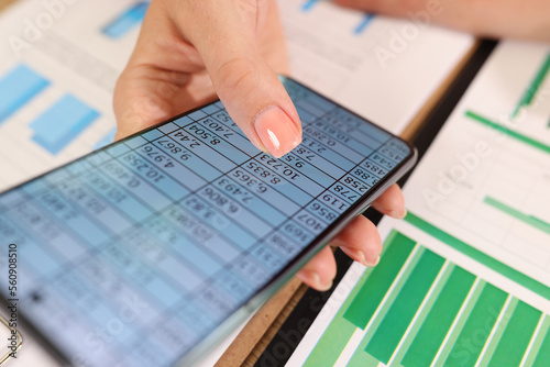 Close up of business woman hand with financial data on smartphone screen and office table with documents.