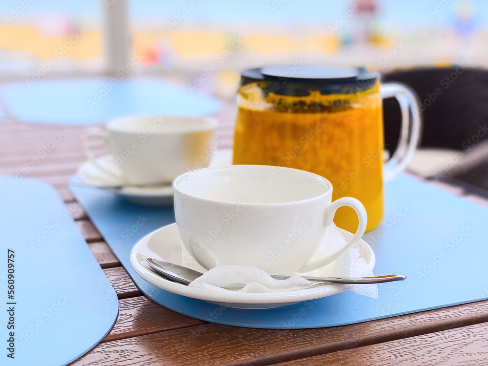 Two white cups and a transparent teapot with sea buckthorn tea on a blue background in an outdoor cafe. Beach, summer. Mobile photography. Horizontal. Two white cups and a transparent teapot with sea buckthorn tea on a blue background in an outdoor cafe. Beach, summer. Mobile photography. Horizontal.