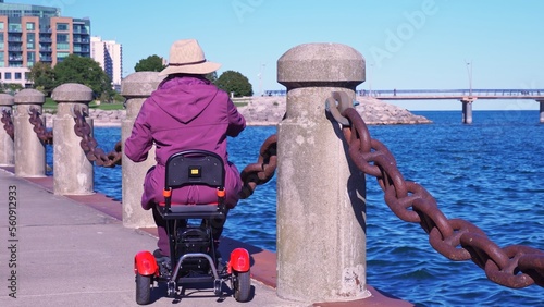 Woman with disability in the wheelchair mobility scooter enjoying park near lake water. Carefree woman with mobility issue uses electric three wheel wheelchair scooter having fun. photo