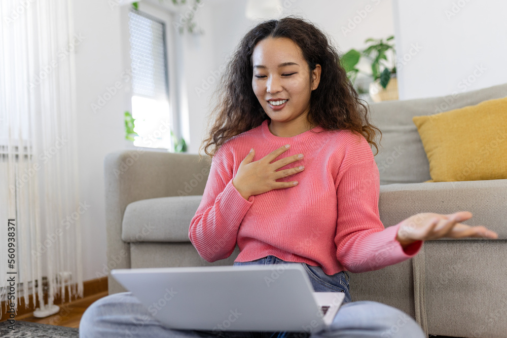 Happy young Asian woman having fun doing video call using laptop in her home, waving hand video conference calling on laptop computer sit on sofa distance learn zoom online virtual meeting at home.