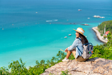 Asian woman with backpack travel at tropical island and resting on mountain peak in summer sunny day. Attractive girl enjoy outdoor lifestyle looking beautiful ocean nature on beach holiday vacation.