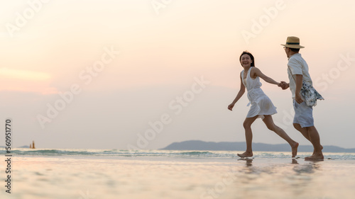 Happy Asian family couple holding hands walking and playing together on the beach at summer sunset. Husband and wife enjoy and fun outdoor lifestyle travel tropical island beach on holiday vacation.