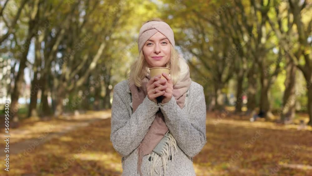 Front view of attractive girl wearing stylish clothes, walking in park. Beautiful, blonde female looking at camera, smiling, warming hands with coffee, Concept of autumn and coziness. Front view of attractive girl wearing stylish clothes, walking in park. Beautiful, blonde female looking at camera, smiling, warming hands with coffee, Concept of autumn and coziness.