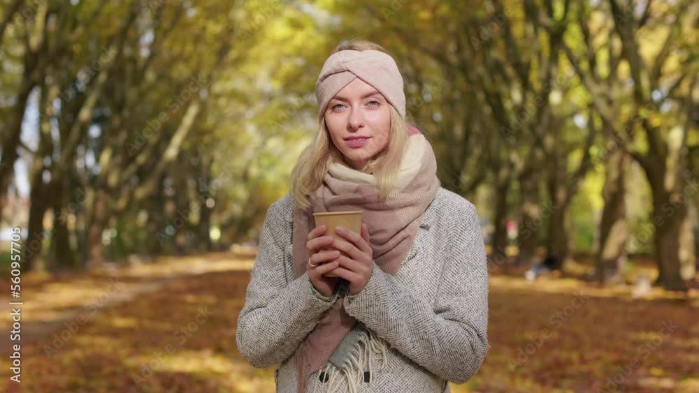 Front view of attractive, blonde woman walking in park with golden trees. Beautiful, young lady standing, looking at camera, holding paper cup. Concept of sincere emotions. Front view of attractive, blonde woman walking in park with golden trees. Beautiful, young lady standing, looking at camera, holding paper cup. Concept of sincere emotions.