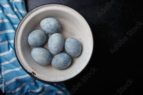 Easter eggs painted blue in white bowl on black wooden background with striped fabric. Top view, copy space