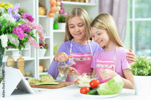 Two girls in pink aprons preparing salad on kitchen with tablet