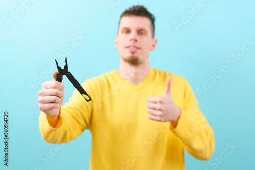 A man holds a multitool and pliers in one device and shows his thumb up on a blue background. Pocket. Black. Blade. Equipment. Metal. Multipurpose. Utility. Survival. Open. Folding. Object. Handle