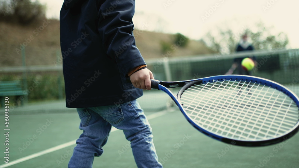 Back of kid playing tennis with parent during winter season. Child wearing coat and beanie hitting ball. Small boy plays sport outdoors
