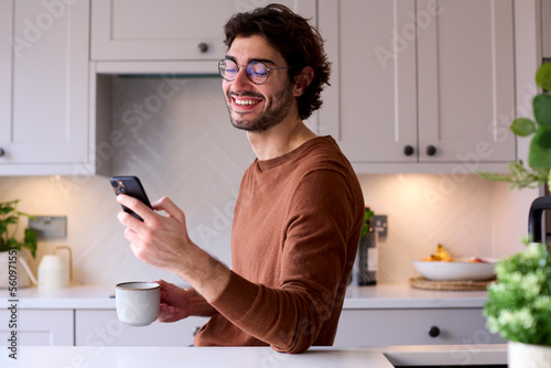 Young Man Relaxing At Home In Kitchen Holding Hot Drink Using Mobile Phone