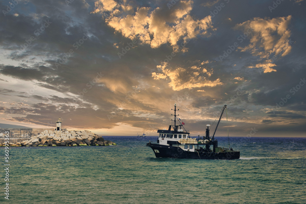 View of a fishing vessel returning from the sea to land and a view of the small harbor, lighthouse and gorgeous dramatic sky.