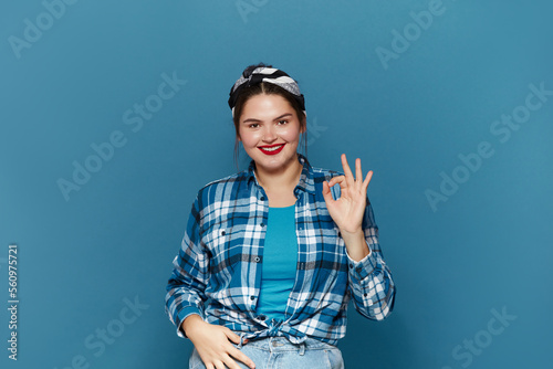 Smiling Woman Ok Gesture Isolated. Portrait of Satisfied Plus Size Girl Standing, Looking at Camera and Showing Ok Sign Gesture. Indoor Studio Shot Isolated on Blue Background 
