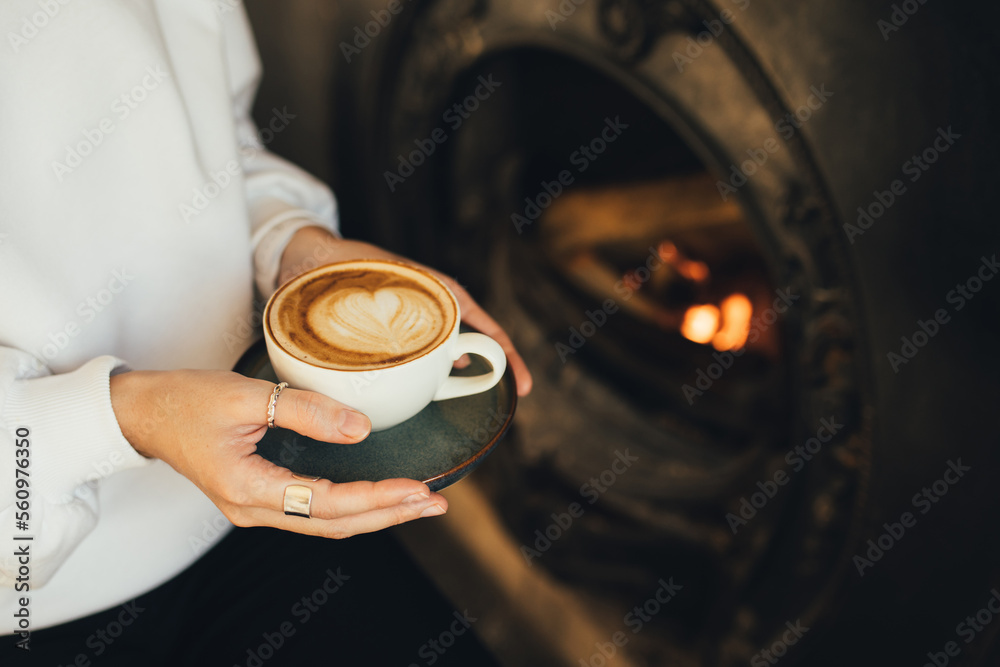 Close up female hands holding cup of hot cappuccino on wooden stool near burning fireplace.