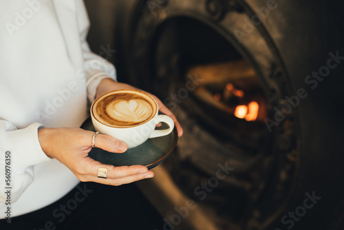 Close up female hands holding cup of hot cappuccino on wooden stool near burning fireplace.