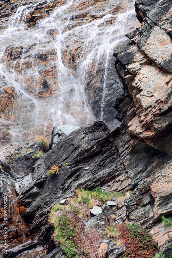 Alpine flowing waterfall washes various color karst overlays of granite rocks, scarce multi-colored rock vegetation nestled in open areas of wet rock, vertical shot, Aosta valley, Italy Alpine flowing waterfall washes various color karst overlays of granite rocks, scarce multi-colored rock vegetation nestled in open areas of wet rock, vertical shot, Aosta valley, Italy