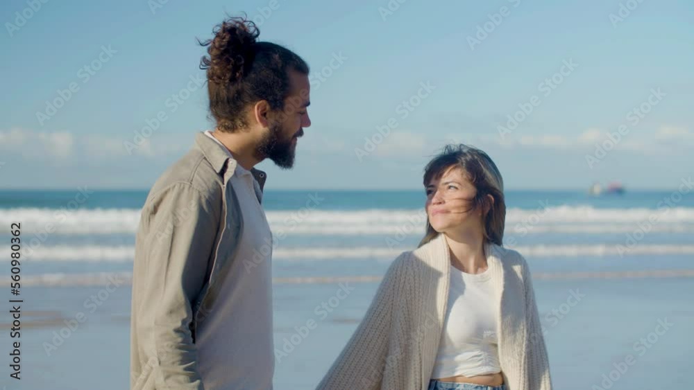 Beautiful Caucasian couple strolling along seashore while holding hands, smiling and talking. Handsome guy with beard and man bun spending time with charming young lady outside. Love, travel concept Beautiful Caucasian couple strolling along seashore while holding hands, smiling and talking. Handsome guy with beard and man bun spending time with charming young lady outside. Love, travel concept