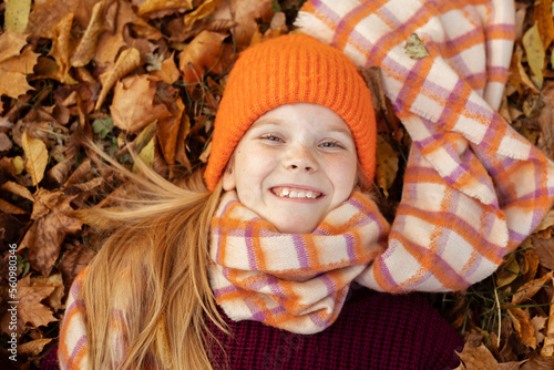 Top view of awesome teenage girl child wearing vinous sweater, orange hat, scarf, lying on yellow fallen maple leaves.