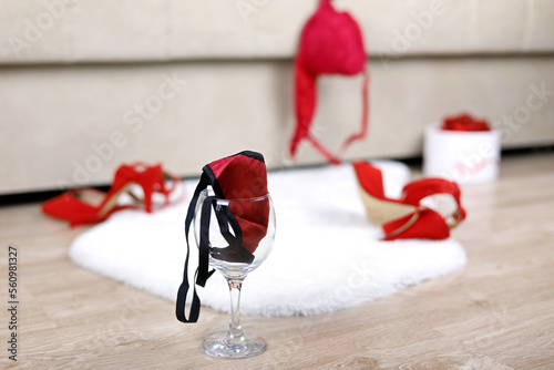 Wine glass with female thong panties, removed red bra and shoes on a floor near bed. Sex concept, romantic night of passion, sexy lingerie
