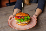 Cropped photo of woman sitting at table, holding big pink plate with fresh burger with lettuce, onion, tomato, chicken.