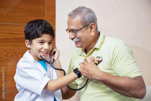 Playful indian boy kid wearing doctor uniform dress standing doing check heartbeat of with his grandfather with stethoscope at home. Closeup