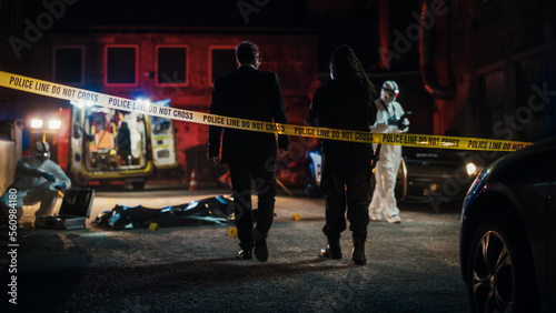 Cinematic Shot: Lieutenant Arriving At a Crime Scene, Crossing the Yellow Tape, Listening to Briefing from First Responder Officer. Detective Checking the Body Bag. Forensics Team Gathering Evidence photo