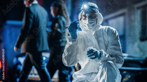 Forensics Technician in Coveralls Inspecting Potential Incriminating Evidence on Crime Scene. He Picks up a Marked Bullet Shell Clue and put it in Plastic Bag for Reservation. Police Gathering Proofs photo