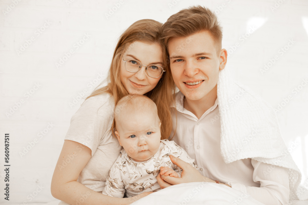 Happy young family of three people sit together and embrace, white background, free copy space. Home family photo of mom, dad and infant child. Concept of parental affection and caring for children