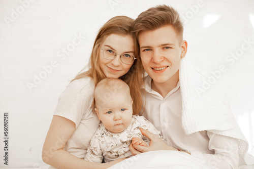 Happy young family of three people sit together and embrace, white background, free copy space. Home family photo of mom, dad and infant child. Concept of parental affection and caring for children