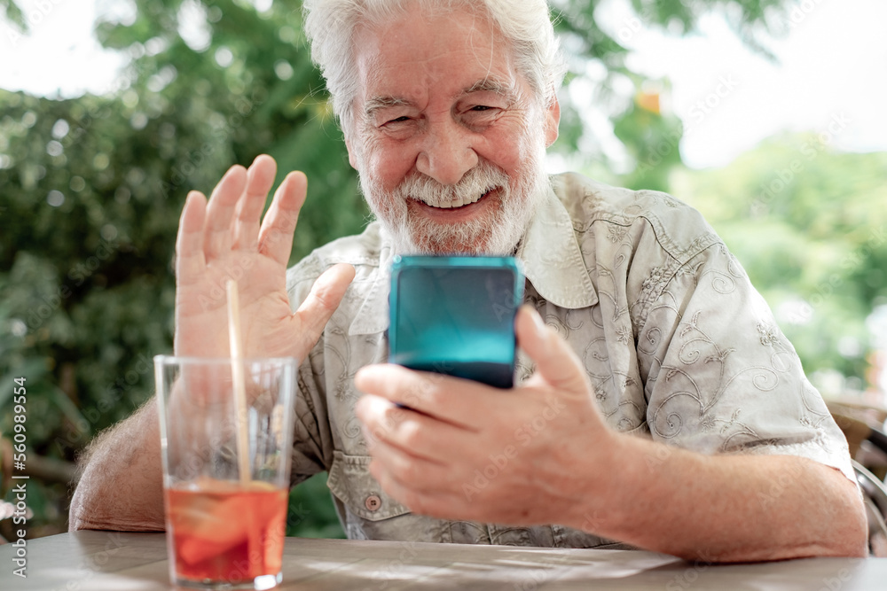Handsome bearded 70s man sitting outdoor at cafe table video chatting by mobile phone. Senior caucasian male using cellphone waving hand Handsome bearded 70s man sitting outdoor at cafe table video chatting by mobile phone. Senior caucasian male using cellphone waving hand