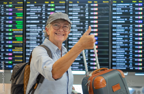 Happy senior woman in airport looking at camera while looking at timetable schedule to check her flight departure gate. Traveler concept people with backpack and suitcases.