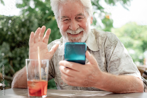 Handsome bearded 70s man sitting outdoor at cafe table video chatting by mobile phone. Senior caucasian male using cellphone waving hand