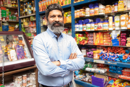 Portrait of happy mature Indian man standing at grocery shop or supermarket with cross arm.