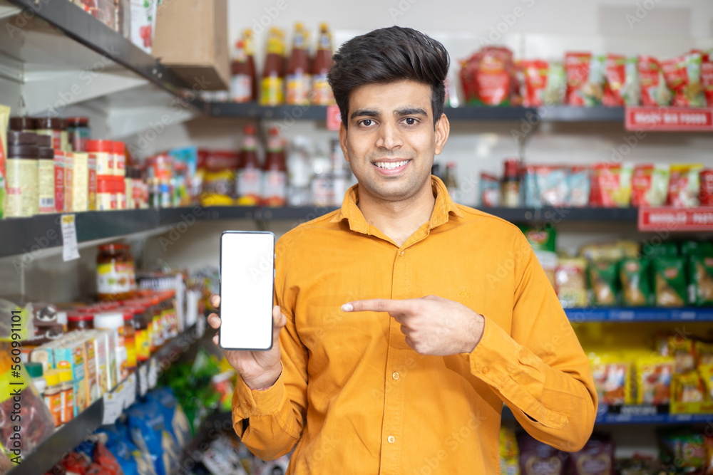Portrait of happy handsome young Indian man showing smart phone with blank display screen to put advertisement at grocery shop or supermarket, Closeup.