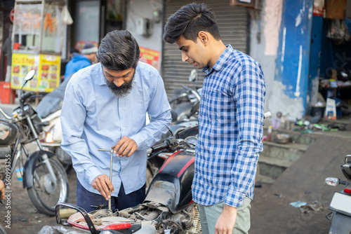 Professional Indian mechanic repairs motorcycle with screwdriver. Asian man repairing motorbike in repair shop along with customer.