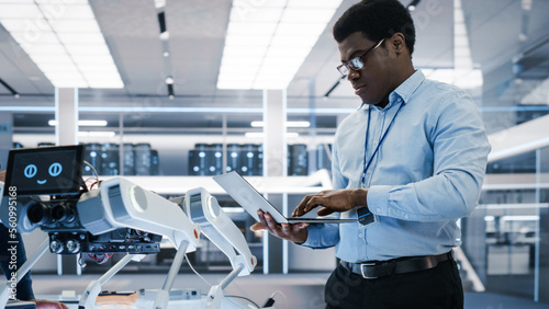 Talented African Robotics Engineer Using Laptop Computer, Analyzing Robotic Machine Concept in a High Tech Factory. Black Scientist Manipulate and Program the Robot for Work.