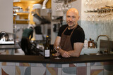 portrait of caucasian barman and owner of a winebar standing behind the counter with a bottle and a glass of wine