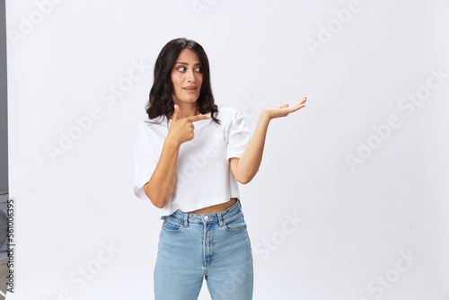 Woman in white t-shirt on white background brunette hands up gestures and signals poses in jeans emotion, lifestyle smiles, copy space