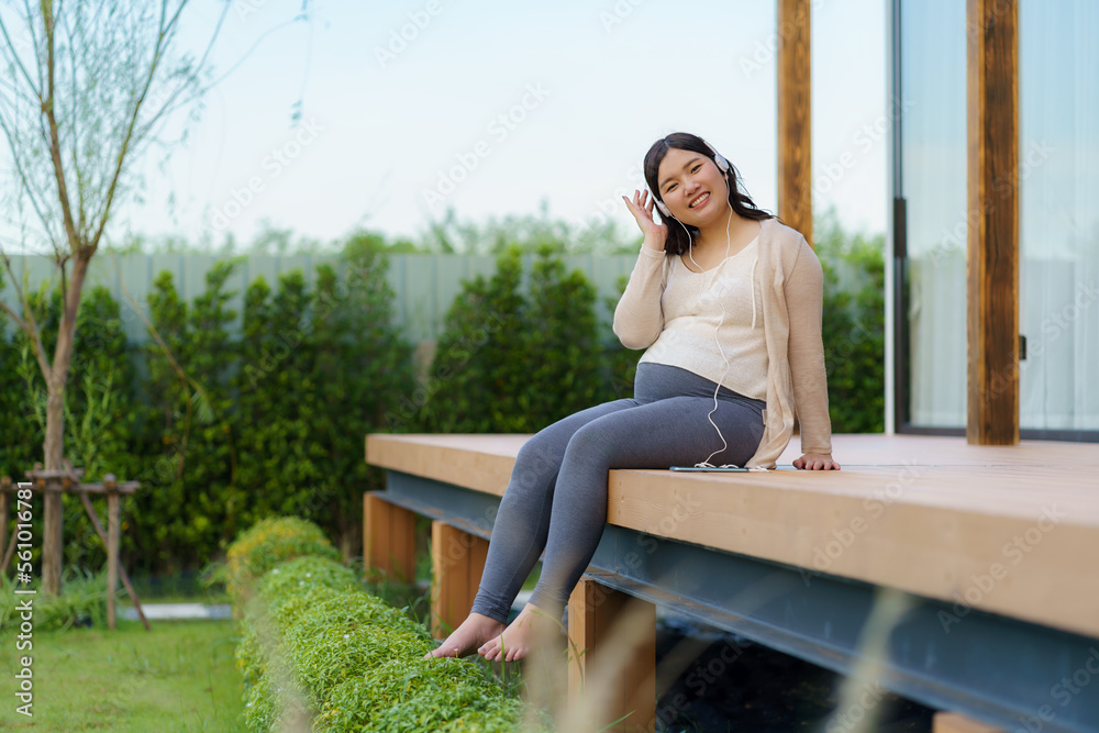 Fototapeta premium Portrait of pregnant Happy Asian woman sitting on terrace at home and using smart phone for listening music in headphones.