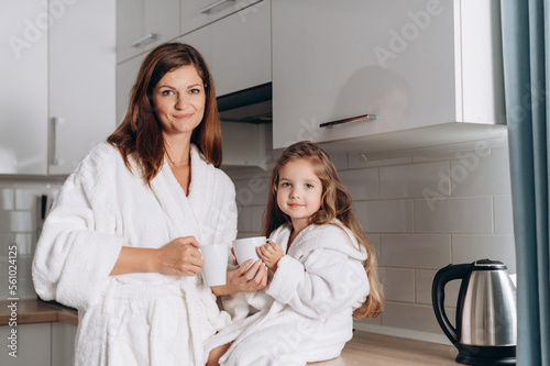 A young mother and her preschool daughter drink tea from mugs in the kitchen. Portrait of mother and daughter at home.