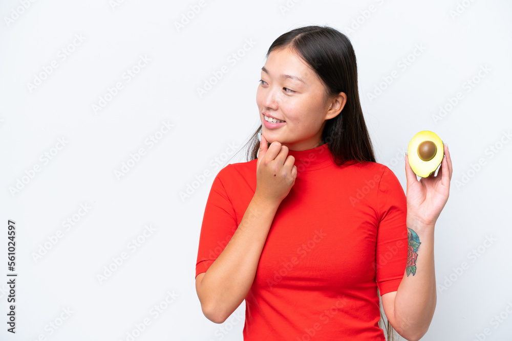 Young Asian woman holding an avocado isolated on white background thinking an idea and looking side Young Asian woman holding an avocado isolated on white background thinking an idea and looking side