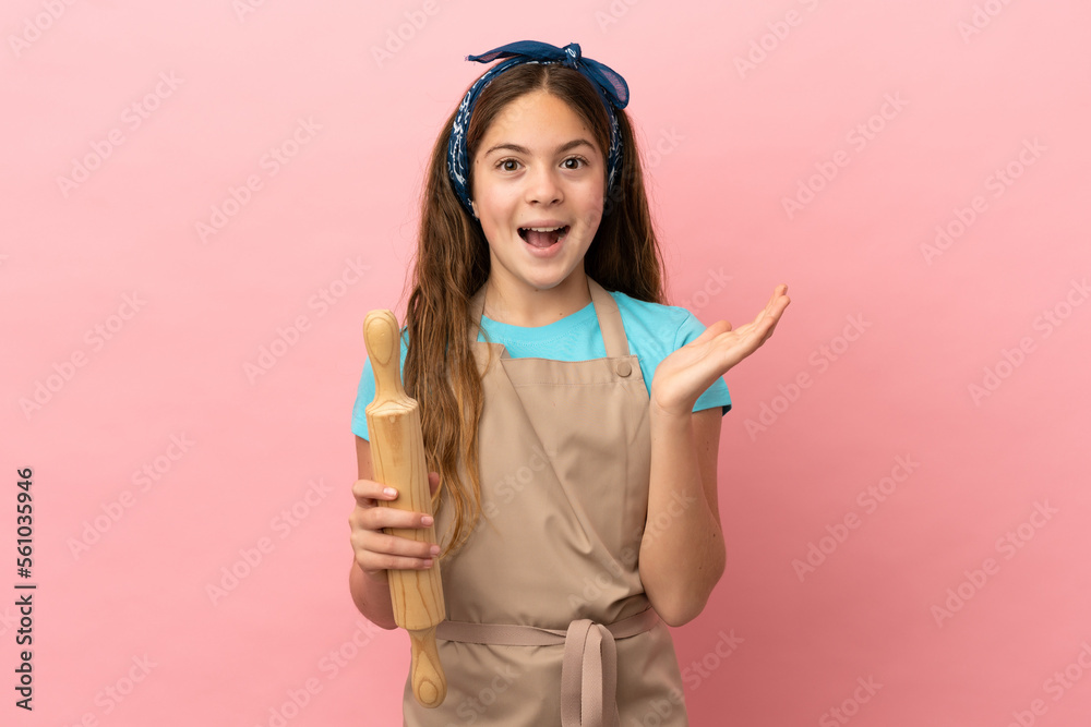 Little caucasian girl holding a rolling pin isolated on pink background with shocked facial expression