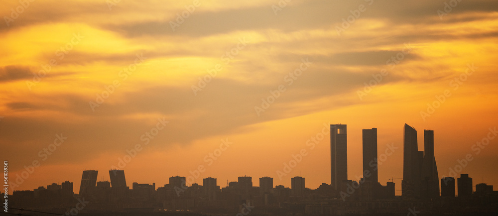 Fototapeta premium Panoramic view where the sun silhouettes the skyscrapers of Madrid's skyline, known as the 'Four Towers Business Area', during sunset