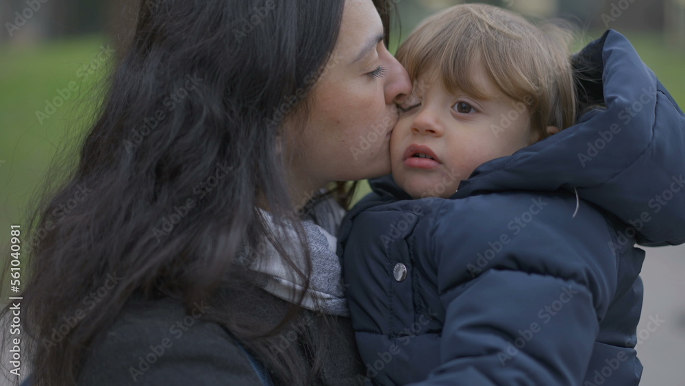 Mother walking outside holding baby toddler in arms, mom kissing son in cheek Mother walking outside holding baby toddler in arms, mom kissing son in cheek
