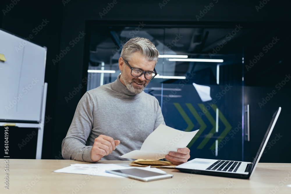 Senior gray-haired man working in office at desk with laptop and documents. an envelope with a letter, bills, and reports, good news. Senior gray-haired man working in office at desk with laptop and documents. an envelope with a letter, bills, and reports, good news.
