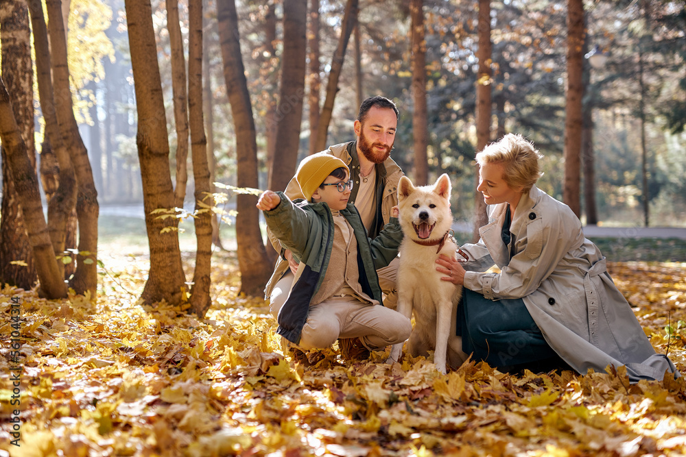 Fototapeta premium happy family with dog on walk in autumn park. family, pet, domestic animal and people concept. young man, woman and child boy in coats outdoors on warm weather. human emotions concept