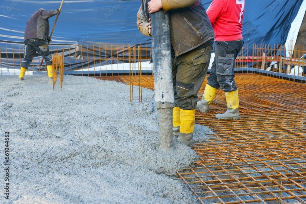 Bauarbeiter gießen die Bodenplatte eines Einfamilienhaus-Neubaus über der Baustahlmatte mit der Betonpumpe, rütteln sie mit Betonrüttler und glätten sie mit Schiebern Bauarbeiter gießen die Bodenplatte eines Einfamilienhaus-Neubaus über der Baustahlmatte mit der Betonpumpe, rütteln sie mit Betonrüttler und glätten sie mit Schiebern