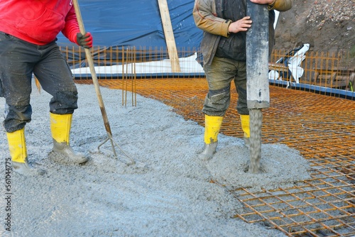 Bauarbeiter gießen die Bodenplatte eines Einfamilienhaus-Neubaus über der Baustahlmatte mit der Betonpumpe, rütteln sie mit Betonrüttler und glätten sie mit Schiebern