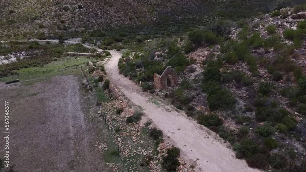 Tracking shot flying forwards and down over the ruins of a small house next to a road, a car drives into the shot, in Cloetesberg, Western Cape, South Africa. Tracking shot flying forwards and down over the ruins of a small house next to a road, a car drives into the shot, in Cloetesberg, Western Cape, South Africa.